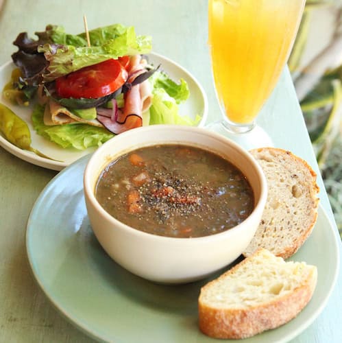 White bowl of soup with bread slices on white plate and salad on another plate and glass of yellow drink