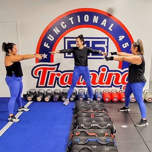 Three woman wearing black shirts and blue pants pointing at each other in front of mural at F45 Training Gym