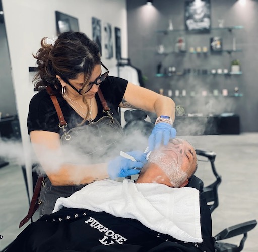 Purpose LA Barbershop woman shaving a man's beard while steam blows on his face