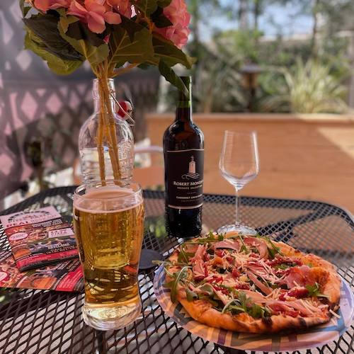 Pizza on an outdoor table with a beer and bottle of wine and menu