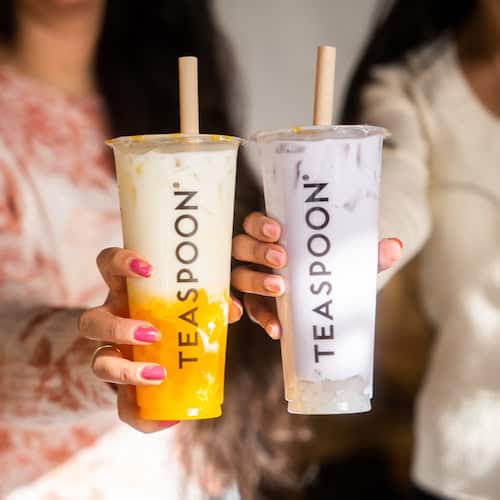 Two women with brown hair holding out two Teaspoon Boba drinks one orange and one purple