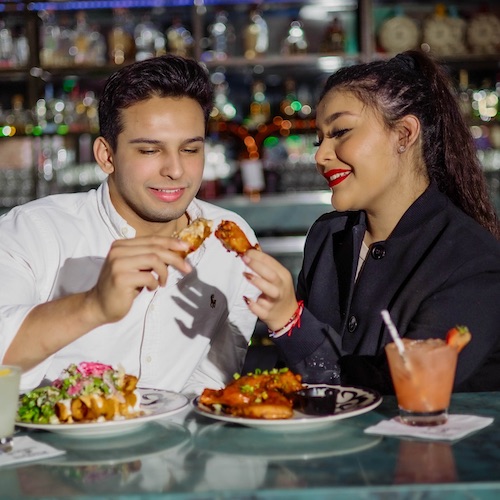 A couple shares two plates of Mexican food and a pink drink at a bar