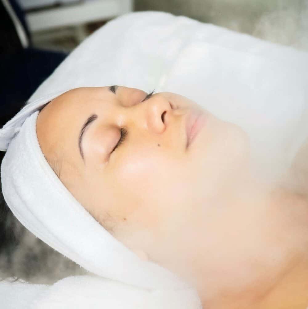 Woman receiving a facial steam treatment at a spa, with eyes closed and wrapped in a white towel, creating a calm and serene atmosphere.