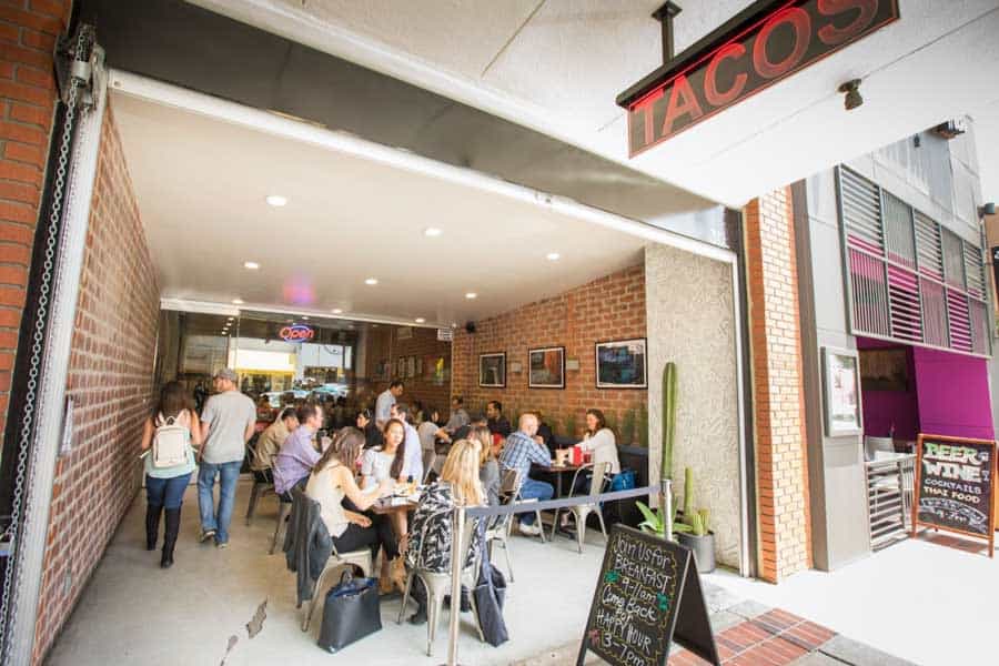 Street view of a busy taco restaurant with an open entrance, where customers are seated inside enjoying meals. A sign above reads 'Tacos,' and a sidewalk board advertises specials.