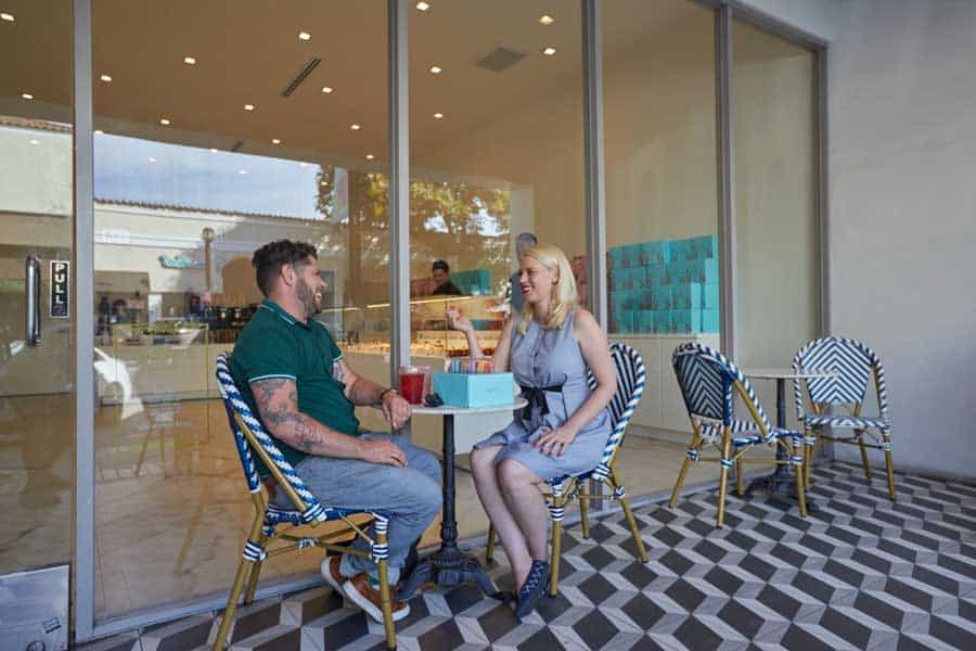 A man and woman sit at an outdoor café table, engaged in conversation, with a bakery visible behind them through large glass windows. The setting has patterned floor tiles and relaxed seating.