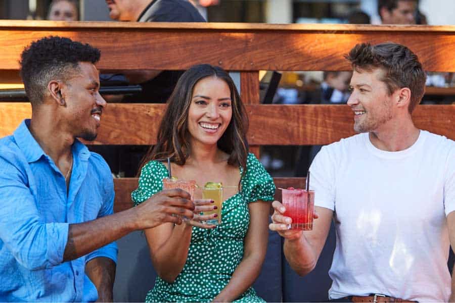 Three friends sitting outdoors, smiling and raising their drinks in a toast, enjoying a casual and lively atmosphere.