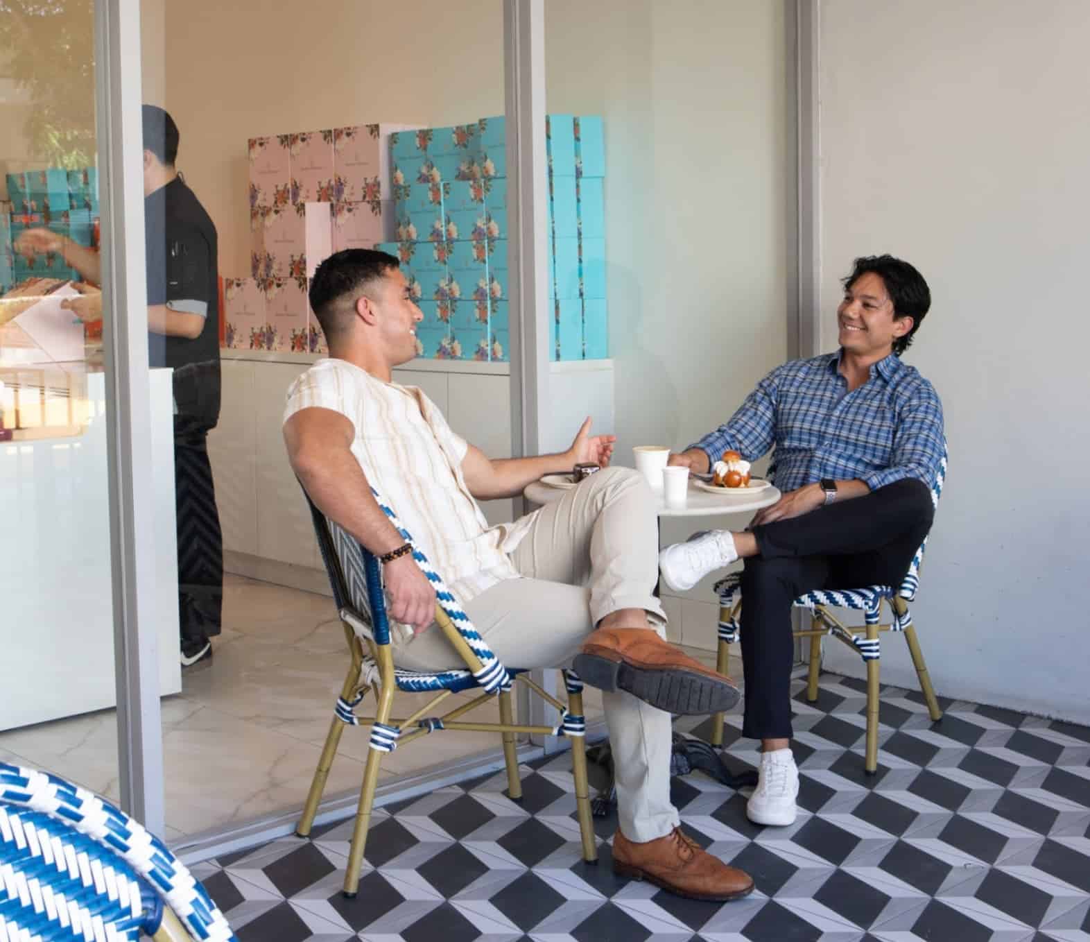 Two men enjoying coffee and pastries while chatting outside Artelice Patisserie, seated on patterned flooring.