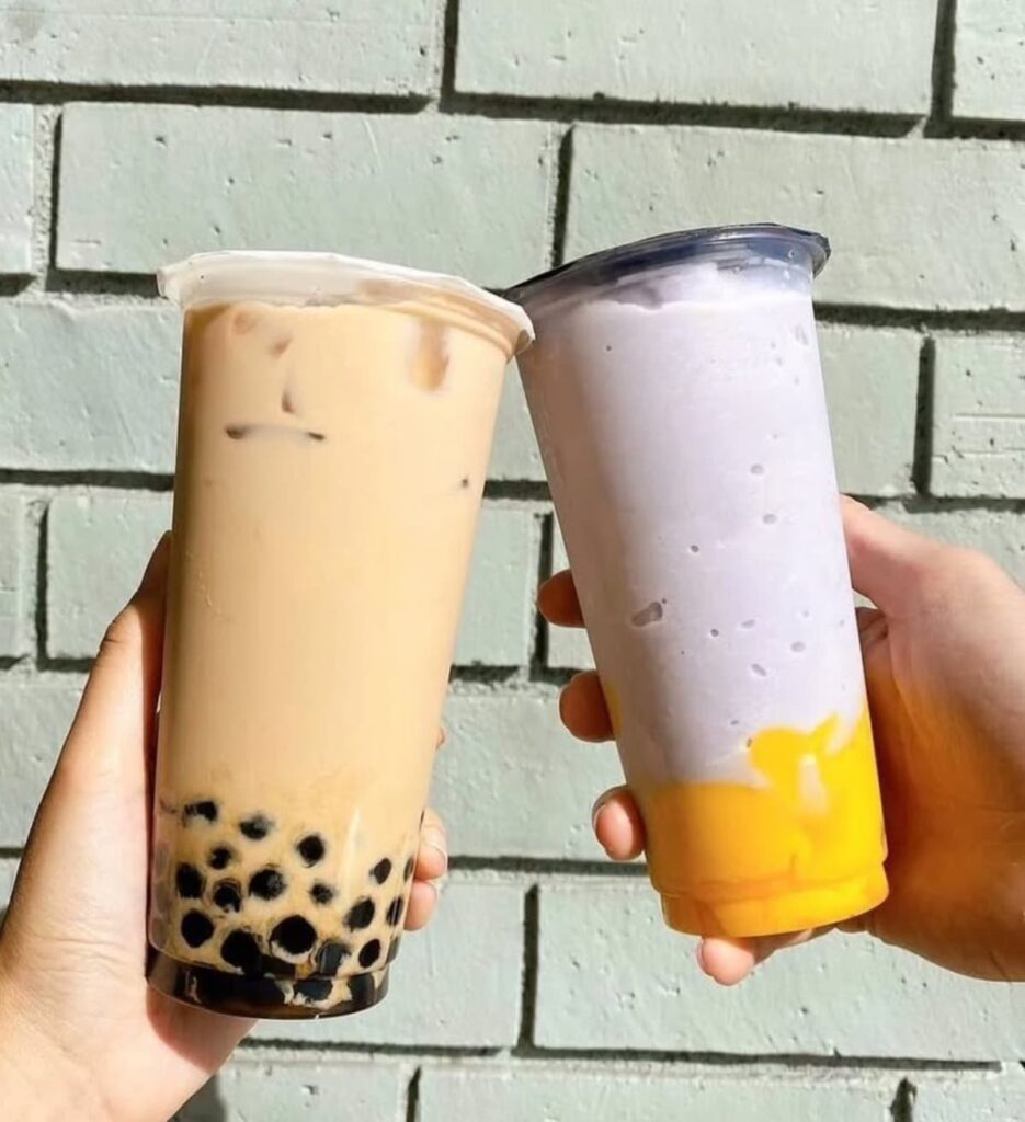 Two hands holding up boba drinks—one with black tapioca pearls and one with purple and yellow swirls—in front of a green brick wall.