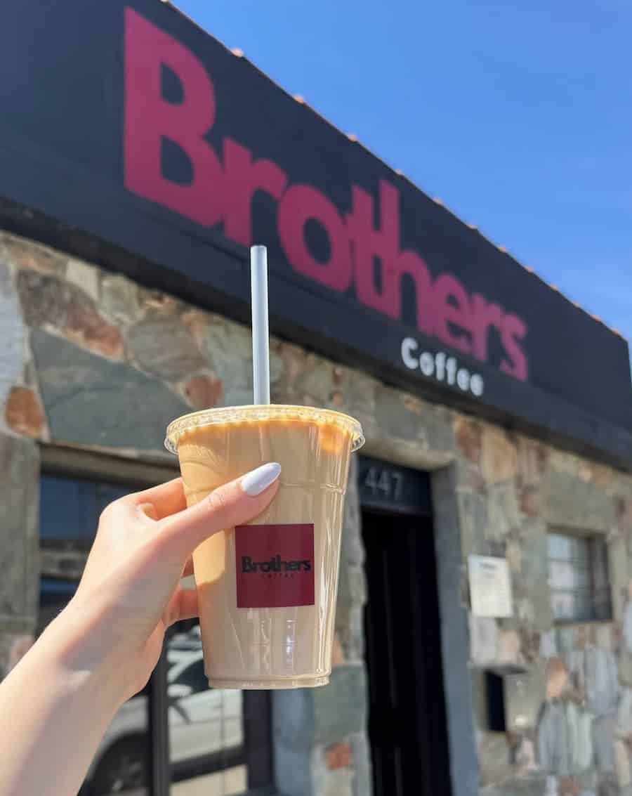 Hand holding an iced coffee in front of the Brothers Coffee storefront with a rustic stone exterior.