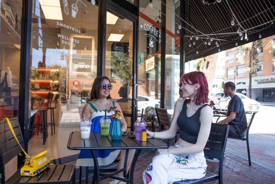 Two women sitting outside Cafe Grano enjoying colorful drinks, laughing and chatting on a sunny day.