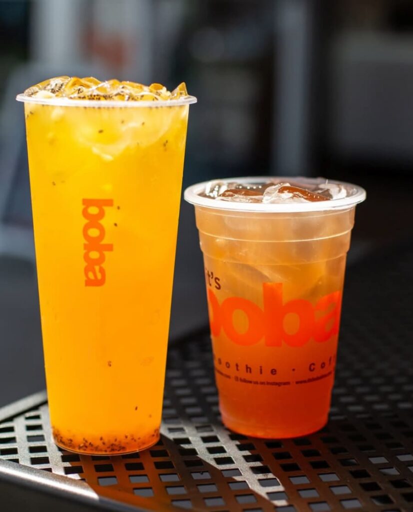 Two iced drinks in plastic cups from It’s Boba Time, placed on a black metal table, with bright orange and amber colors.