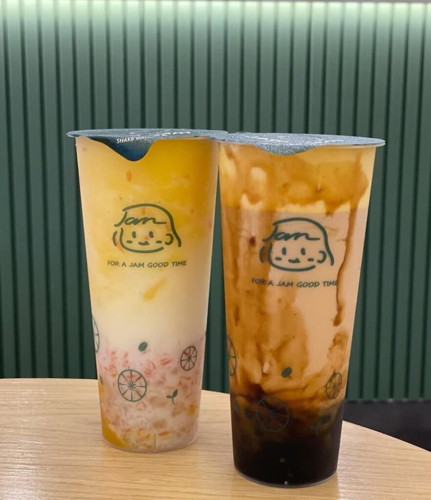 Two colorful bubble tea drinks from Jam Tea Lab with illustrated cup designs, set on a wooden table with a green backdrop.