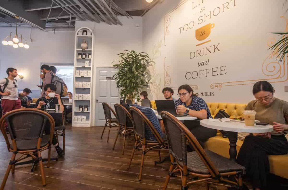People working and relaxing inside a modern coffee shop with the phrase "Life is too short to drink bad coffee" on the wall.