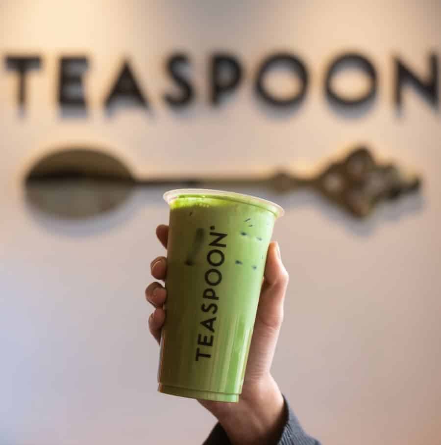 Hand holding a plastic cup of iced matcha tea with the Teaspoon logo, in front of the Teaspoon store wall.