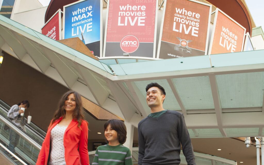A happy family of three — a woman, a child, and a man — walking down escalators in Downtown Burbank with bright banners overhead