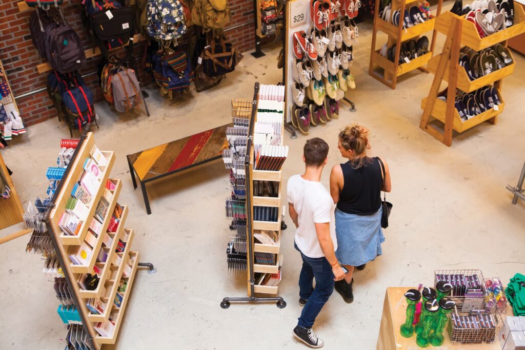 Shoppers browsing through racks of backpacks, shoes, and books inside a store with a warm, inviting atmosphere.