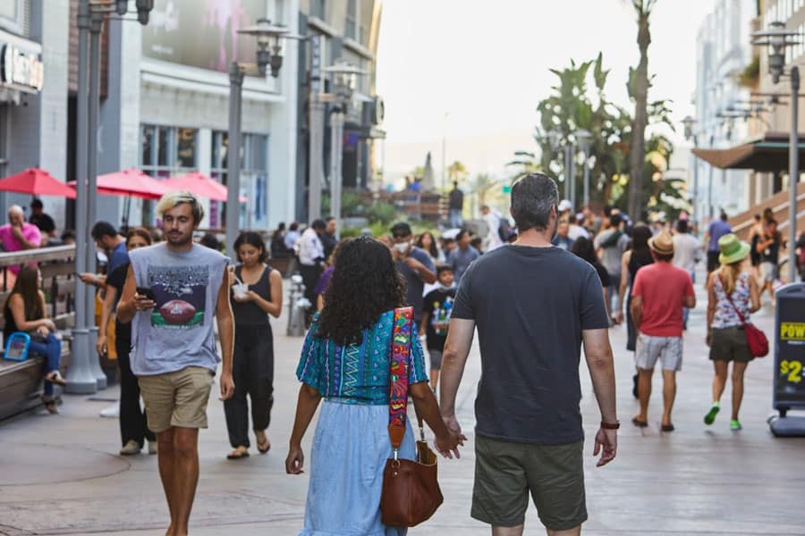 Crowded outdoor shopping street with people walking, dining, and enjoying the atmosphere under umbrellas and palm trees.
