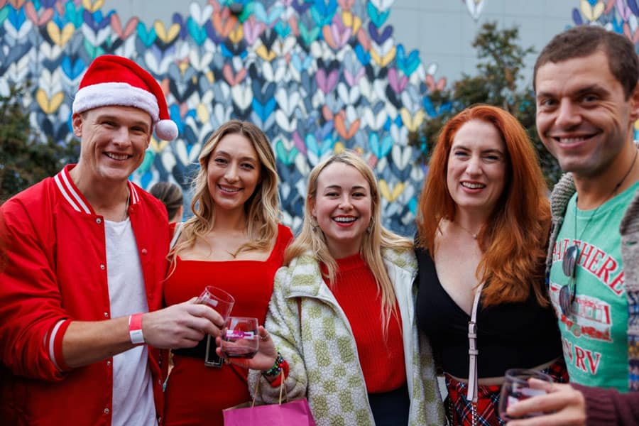 Group of friends posing for a photo at a festive outdoor event, with one person wearing a Santa hat and a colorful mural of hearts in the background.