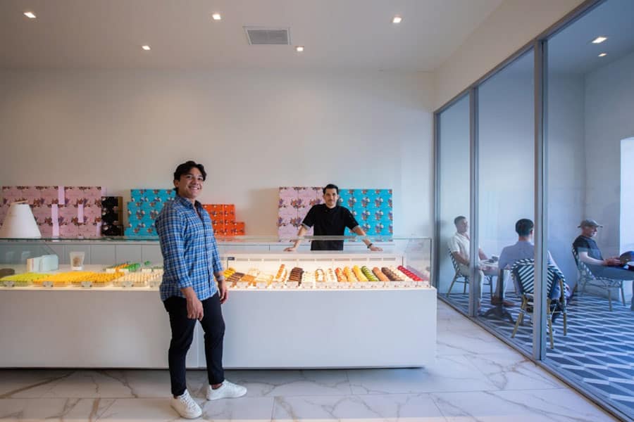 Modern dessert shop interior with display cases of colorful pastries, a smiling staff member in front, and customers seated in a glass-walled side area.