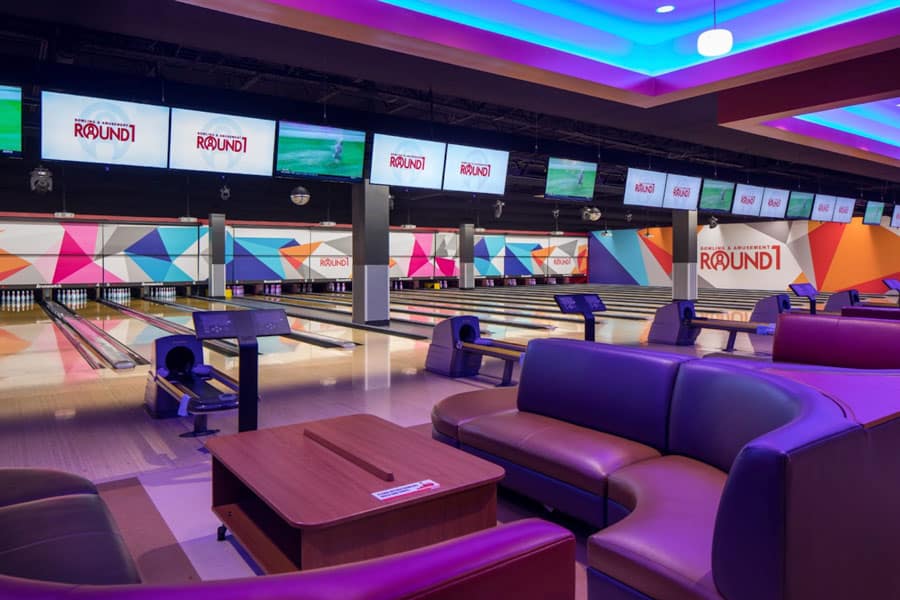 Modern bowling alley interior with colorful geometric wall designs, large screens above the lanes, and lounge seating.