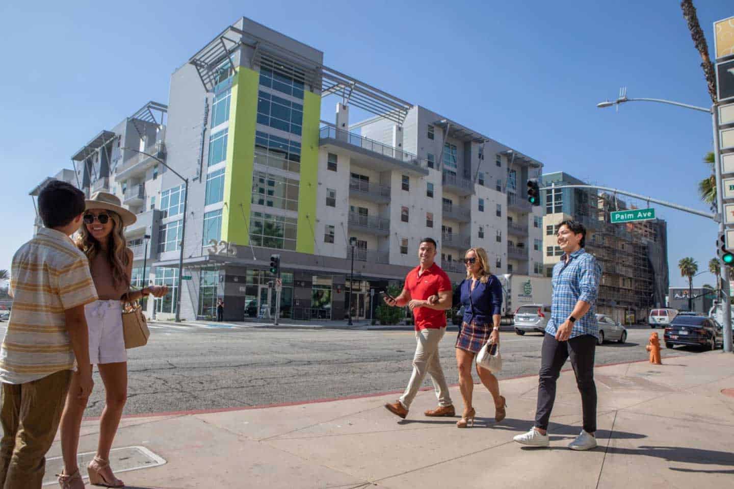 Four people walking together on a sunny city sidewalk, passing a modern apartment building with white and lime green accents, located at the intersection of Palm Ave.