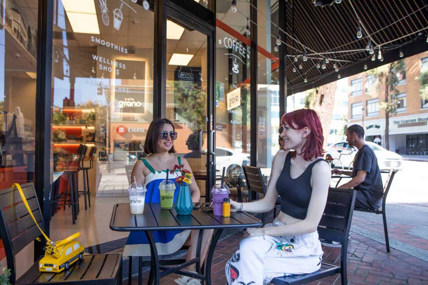 Two young women sit at a black bistro table outside a modern cafe, smiling and talking over colorful smoothies. Large windows behind them display "Smoothies" and "Wellness Shots" text.