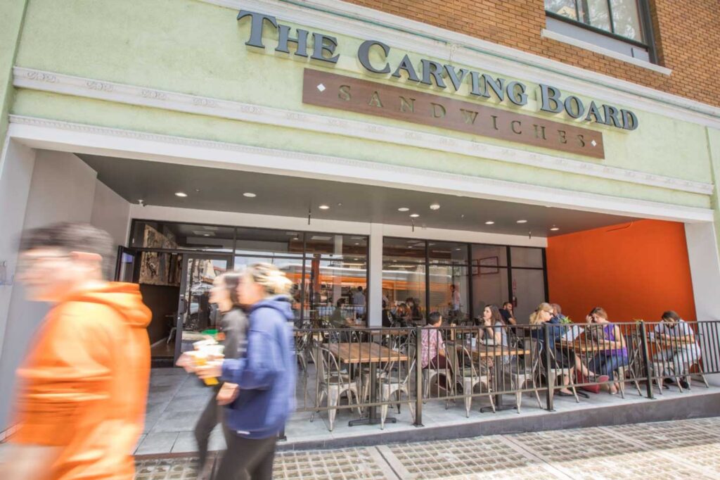 The exterior of "The Carving Board Sandwiches" restaurant, featuring an outdoor patio with metal tables and chairs where several people are dining.