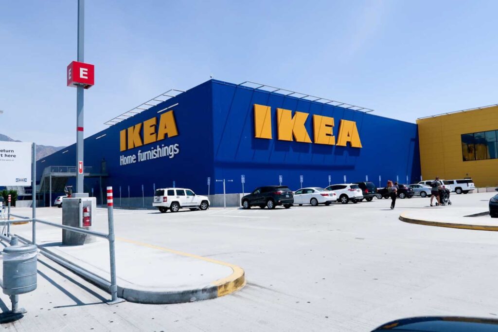 A wide shot of a large blue IKEA home furnishings building with its iconic yellow logo. A bright, sunny parking lot with several cars sits in the foreground.