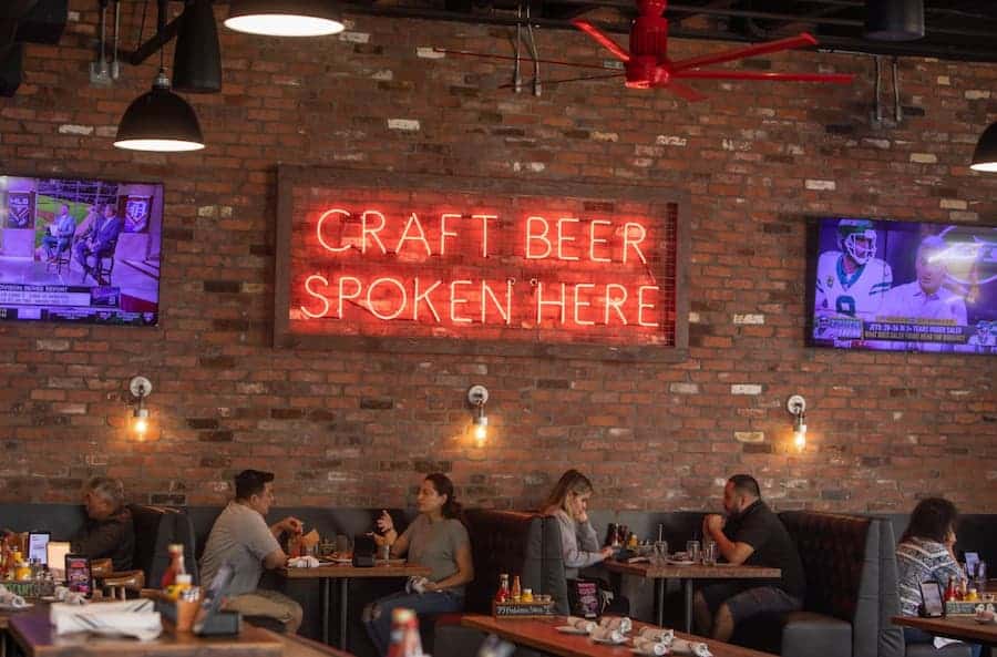 A brick-walled restaurant interior with booths and a neon sign that reads "CRAFT BEER SPOKEN HERE."