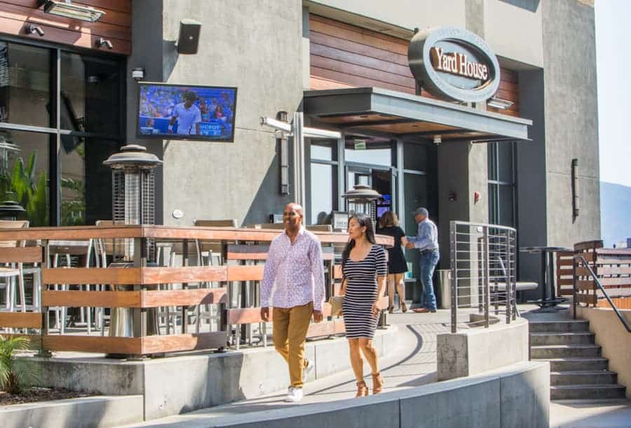A couple walking past the outdoor patio of a Yard House restaurant on a sunny day.