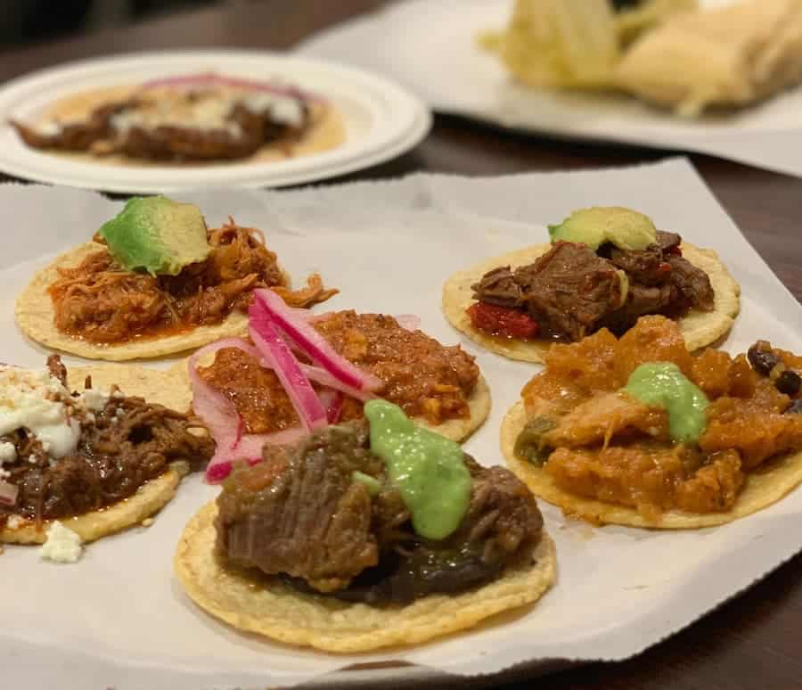 A variety of street tacos on a white plate with toppings like avocado, pickled onions, and salsa.