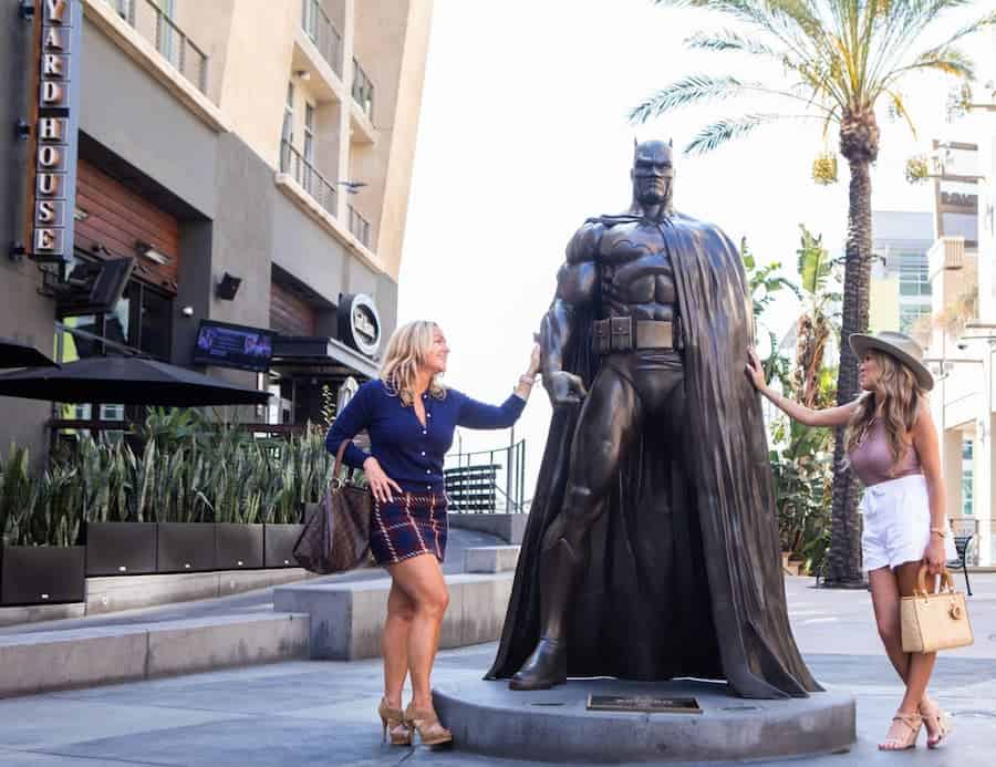 Two women posing and smiling next to a bronze Batman statue in an outdoor shopping area.